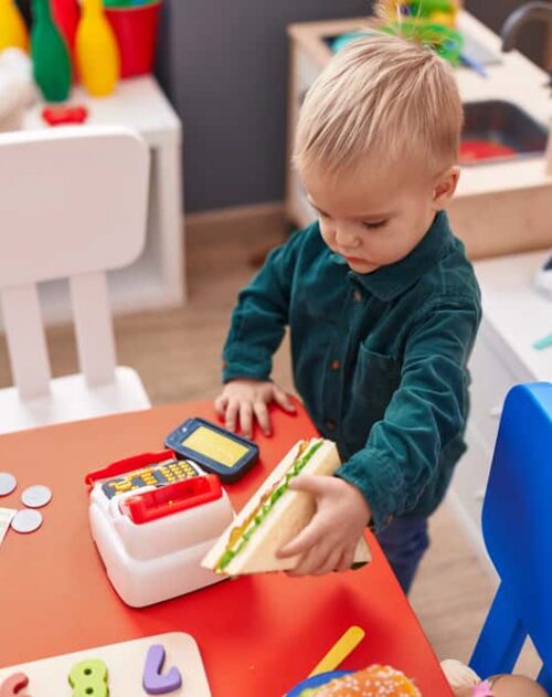 Adorable Caucasian Boy Holding Sandwich And Supermarket Toy At Kindergarten